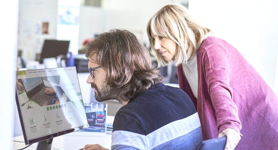 Dos personas delante de un ordenador portátil en la oficina, una con discapacidad visual